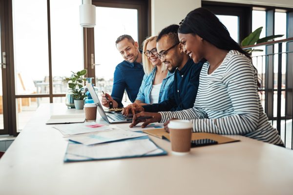 Diverse group of businesspeople smiling while sitting together at a table in an office discussing paperwork and using a laptop