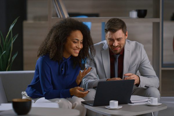 Two multi ethnic employees working together researching laptop in office. Smiling business people video chatting in cafe. Colleagues having online meeting in coworking. Remote work corporate concept.